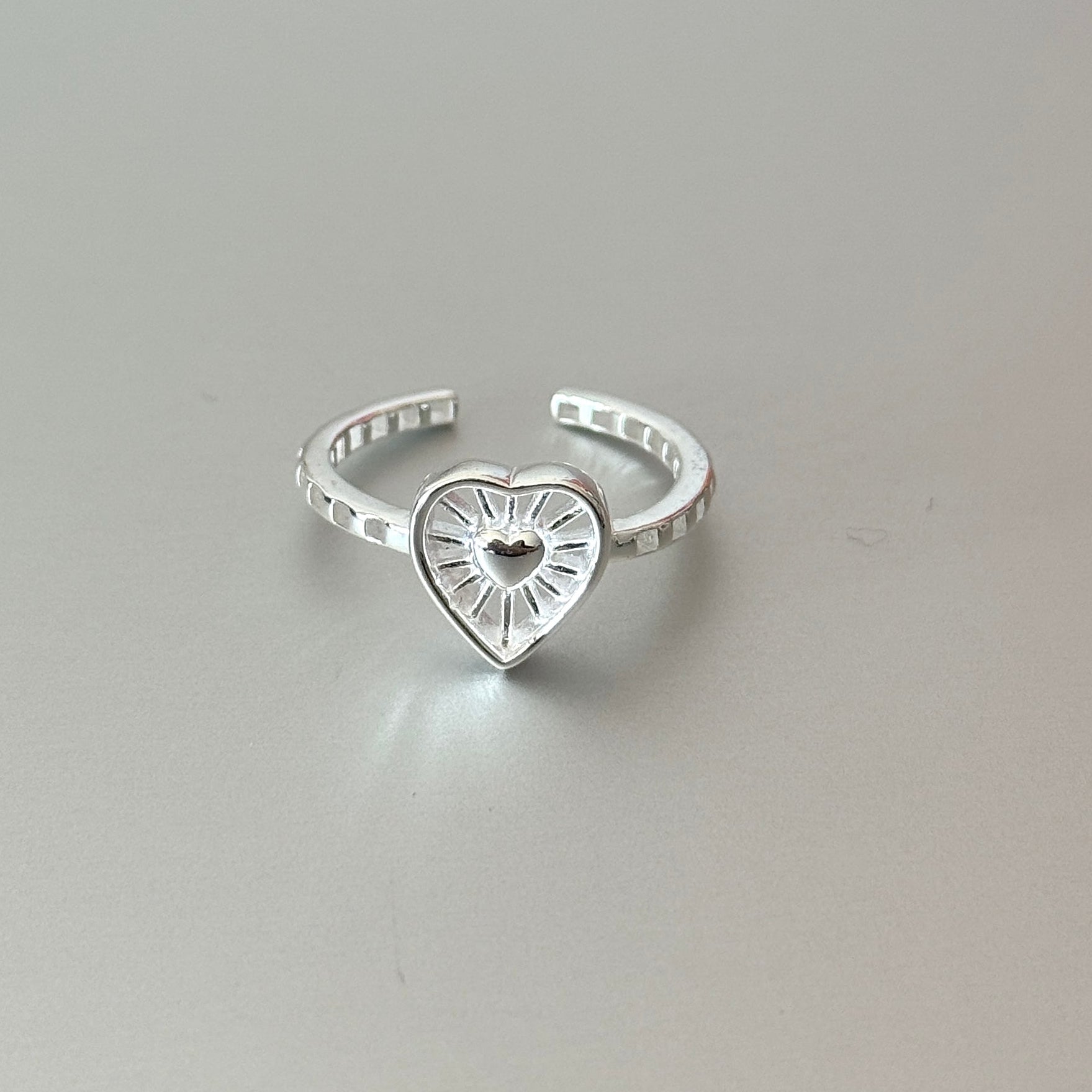 The Thalia open ring in sterling silver, featuring a double heart with sunburst texture, worn on a finger.