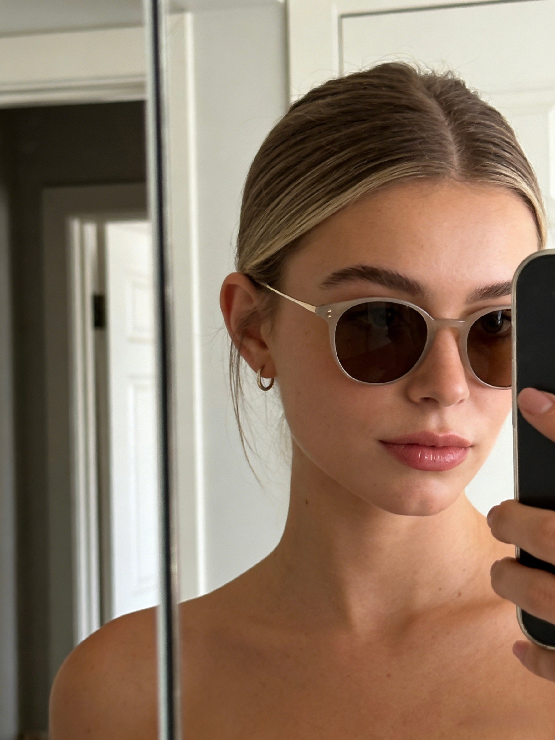Woman wearing sunglasses taking a mirror selfie indoors. Classic round unisex beige color sunglasses | polarised lenses, 100% UV protection, stylish trendy fashion